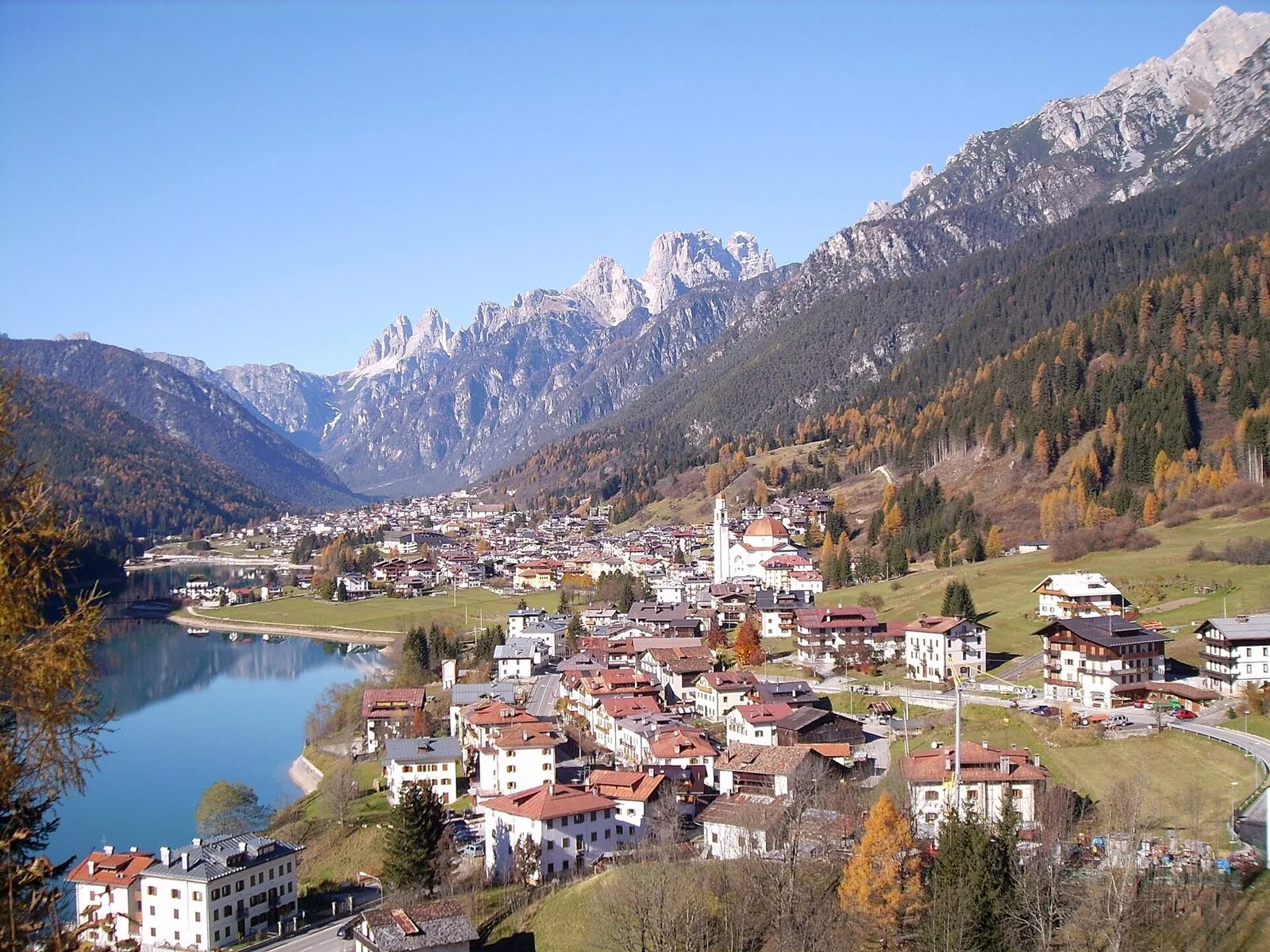 Veduta autunnale di Auronzo di Cadore: le case del paese e il campanile lungo la riva del lago, con larici dorati e le cime dolomitiche sullo sfondo