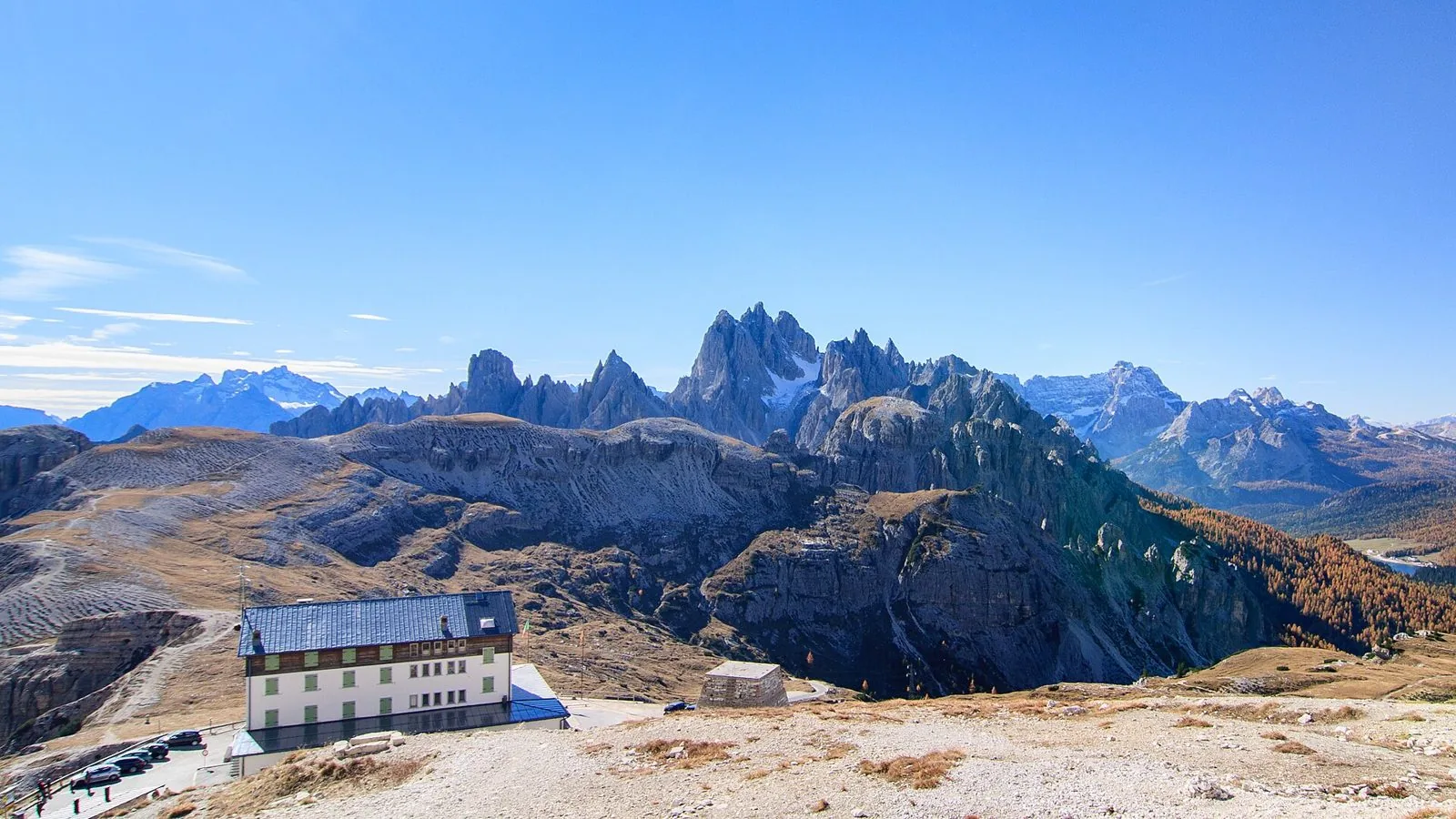 Il Rifugio Auronzo con il parcheggio in primo piano e, sullo sfondo, le guglie dei Cadini di Misurina e i boschi di larici in autunno