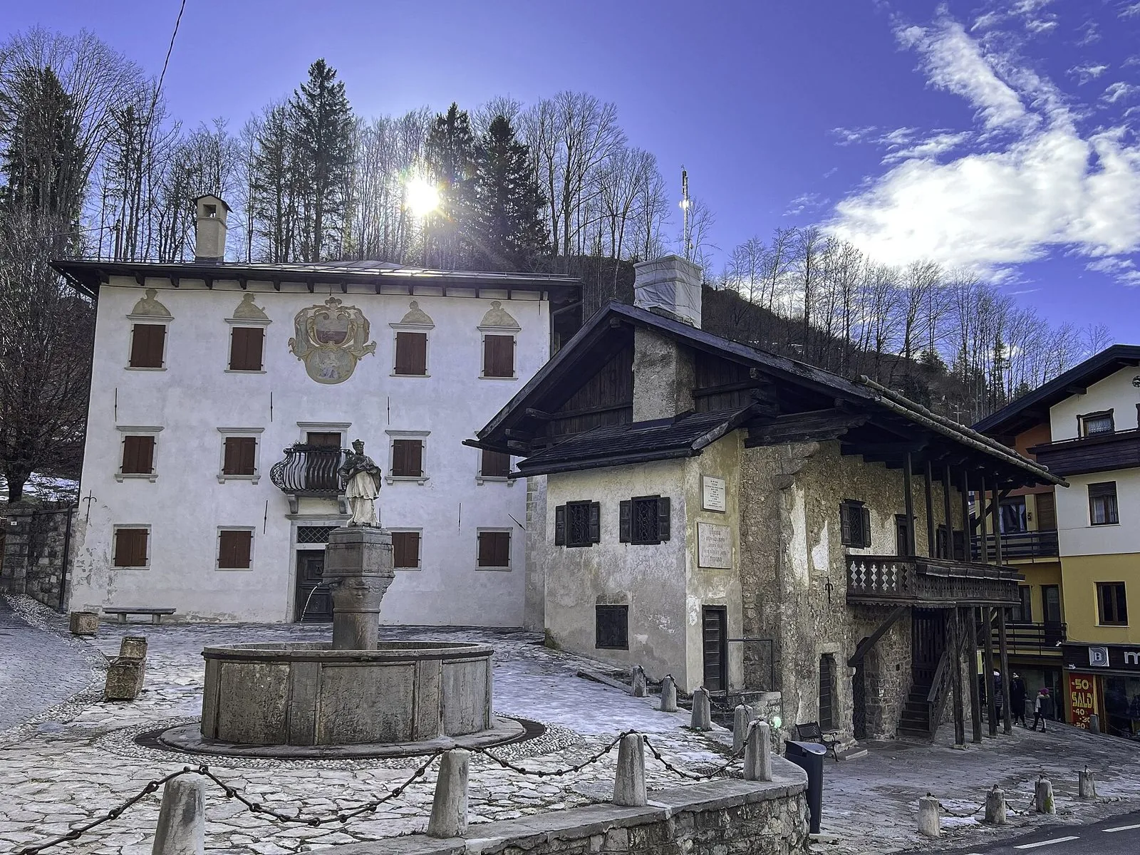 La casa natale di Tiziano a Pieve di Cadore, edificio in pietra con ballatoio e scala esterna in legno, accanto a una fontana con statua e a un palazzo bianco decorato